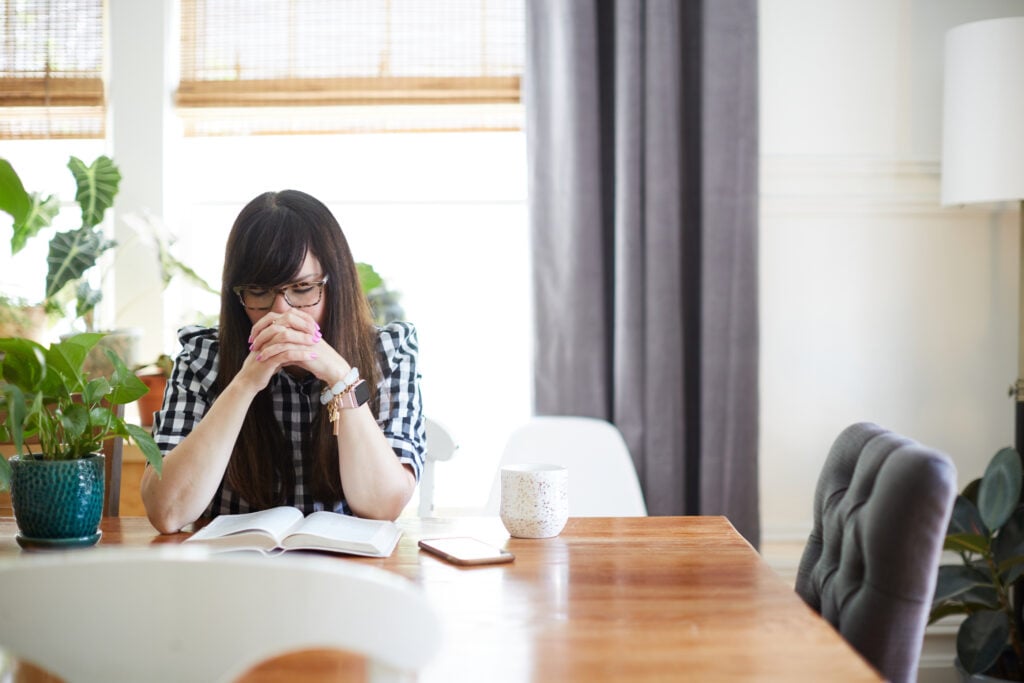 A Christian woman bowing her head and folding her hands in prayer at her dining table with her Bible open in front of her and a mug and her mobile phone beside her