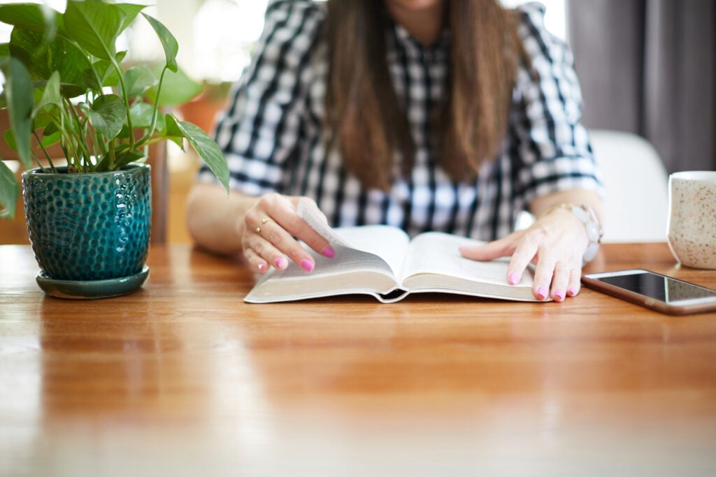A Christian woman reading the Bible at her dining table with a green potted plant, her mobile phone, and a mug beside her
